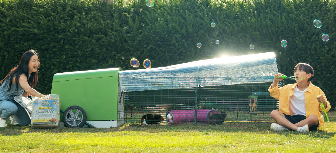 A child blowing bubbles in garden, while his mum is changing the hay inside an Eglu Go Hutch.