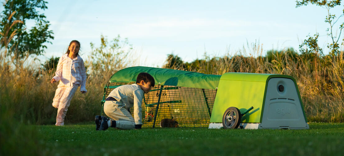 Two children interacting with their guinea pigs next to green Eglu Go hutch in garden.