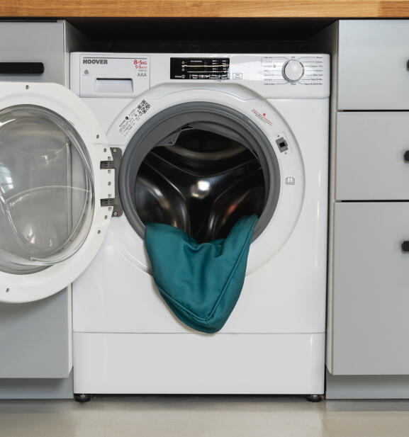 Washing machine with door open and bolster bed cover in shaded spruce print inside.