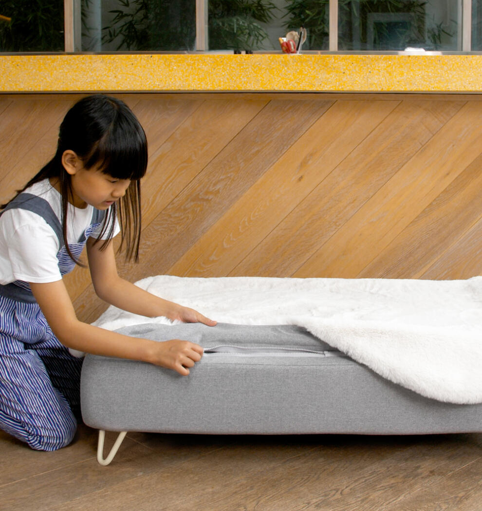Girl zipping a white faux sheepskin topper onto Topology dog bed mattress with white hairpin feet.